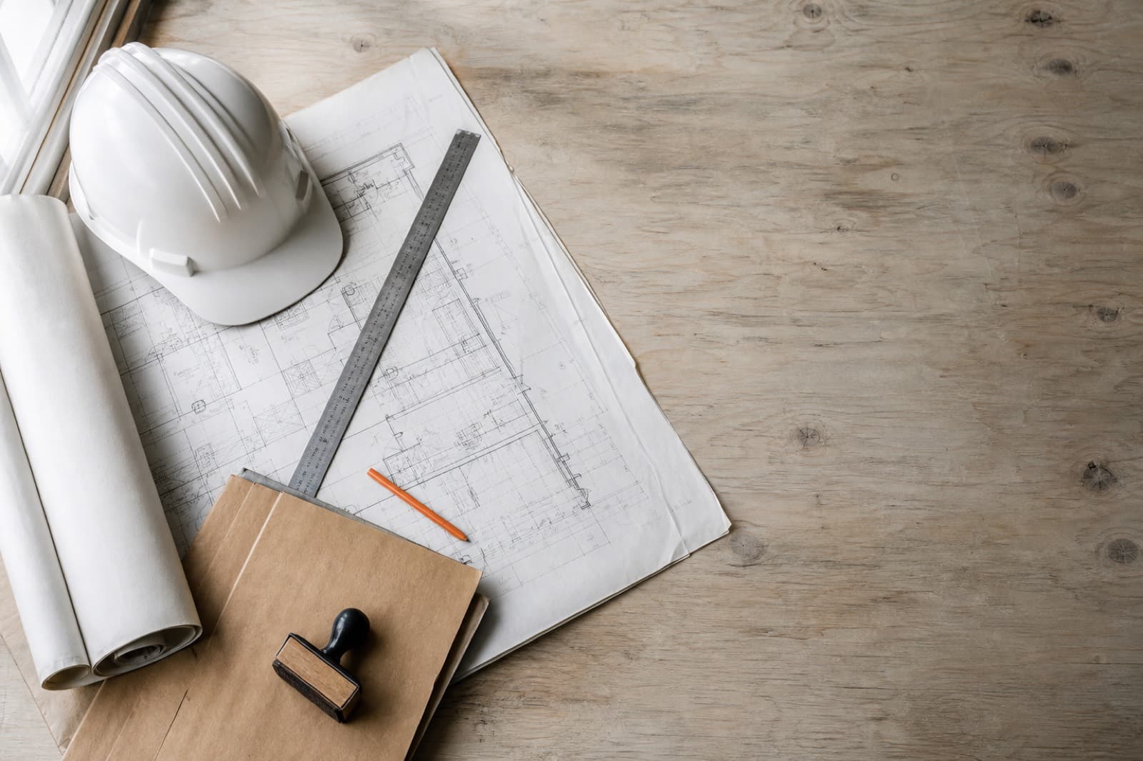A rolled drawing set, hard hat, steel ruler, and stamped permit jacket on a plywood table.
